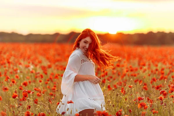 Redhead smiling happy woman in white dress on field of poppies at warm summer sunset