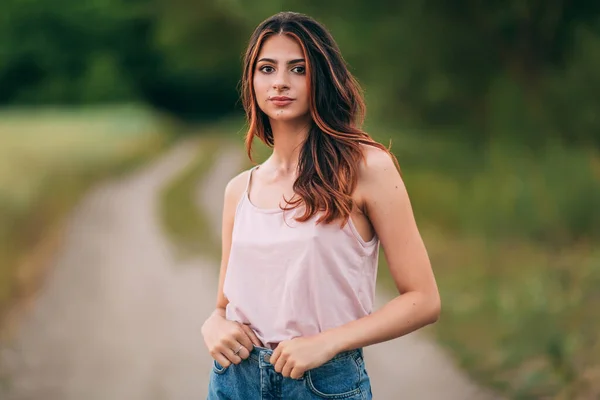 Pembe kolsuz tişörtlü, uzun kahverengi saçlı ve kot şortlu güzel bir genç kadının portresi yaz günbatımında açık havada poz veriyor, şehvetli, ciddi, bulanık bir arka plan.