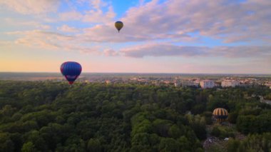 Yaz günbatımında bulutlu mavi gökyüzünde yeşil parkın ve nehrin üzerinde uçan renkli sıcak hava balonlarının insansız hava aracı görüntüsü