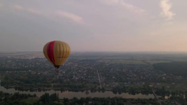 Yeşil parkın üzerinde uçan renkli sıcak hava balonunun ve yaz günbatımında küçük Avrupa kentindeki binaların insansız hava aracı görüntüsü