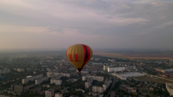 Luftaufnahme eines bunten Heißluftballons, der bei Sonnenuntergang über einem grünen Park und Gebäuden in einer kleinen europäischen Stadt fliegt — Stockvideo