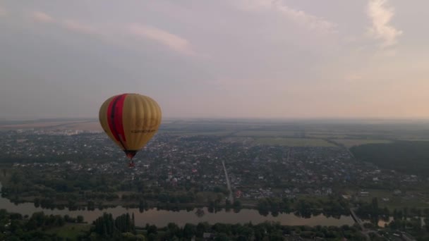 Luftaufnahme eines bunten Heißluftballons, der bei Sonnenuntergang über einem grünen Park und Gebäuden in einer kleinen europäischen Stadt fliegt — Stockvideo