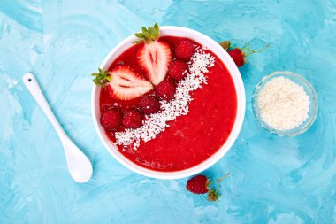 Güler yüzlü kase. Mavi arka plan üzerinde sağlıklı kahvaltı kase. Çilek ve Hindistan cevizi ile ahududu güler yüzlü. Vegan gıda. Düz yatıyordu. Üstten görünüm.