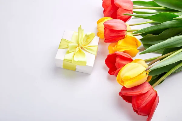White gift box with yellow ribbon near bouquet yellow red tulip on white background. Flat lay. Mother or Woman  Day. Greeting Card. Copy space. Spring.