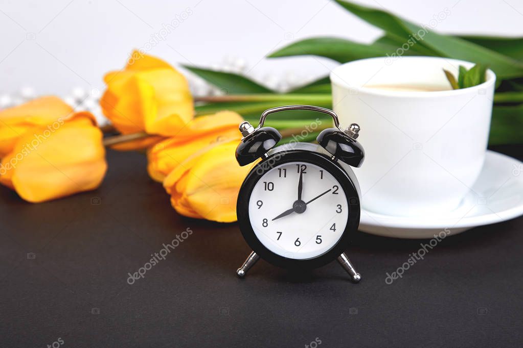 Black alarm clock near bouquet yellow tulips and cup of lemon tea on black background. Mother or Woman  Day. Greeting Card. Good Morning breakfast. Copy space. Spring.