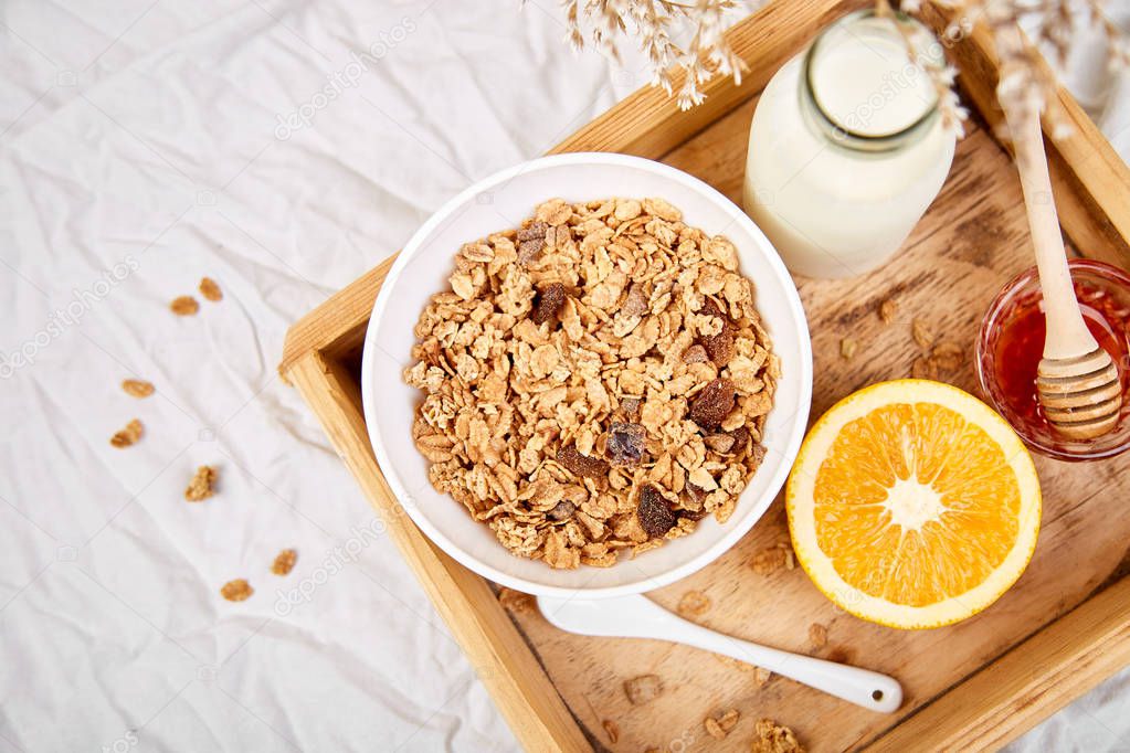 Good morning. Breakfast on white bed sheets. Muesli or granola, milk, orange  on wooden tray from above. Top view.  Flat lay. Copy space. Hotel Room Early Morning.