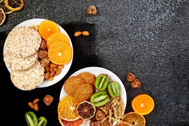 Sağlıklı aperatifler beyaz plakaları - çeşitli yulaf granola bar, pirinç cips, badem, kivi, portakal, siyah arka plan üzerine kurutulmuş. Mix ürün yelpazesine sahip. Üstten Görünüm. Diyet yemekleri. Ücretsiz Gluten. Vegan.