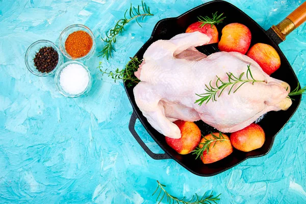 Whole raw chicken in skillet  or iron pan with rosemary leaf, thyme, lemon, red apples on blue background. Ready to cook. Cooking background. Copy space