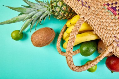 Mavi arka planda organik egzotik meyveler bulunan bir bambu poşeti. Yeniden kullanılabilir bir alışveriş çantasından dökülen taze tropikal meyve çeşitleri. Sağlıklı diyet yemekleri kavramı. Boşluğu kopyala.