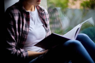 Evdeki pencerenin yanında kitap okuyan güzel bir genç kadın, pencere kenarında oturan bir kız, evde ders çalışan, karantina yapan, evde kalan, yaşam tarzı olan bir kız..