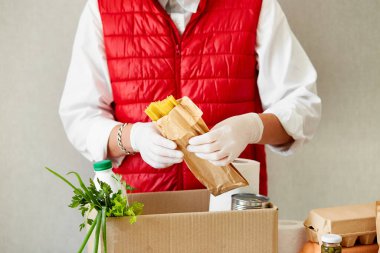 Koruyucu tıbbi maske ve eldiven giyen gönüllü bağış kutusuna yiyecek koyuyor. Kırmızı yelek giyen teslimatçı eleman yiyecek dolu bir kutuyla. Servis karantina pandemik koronavirüsü.