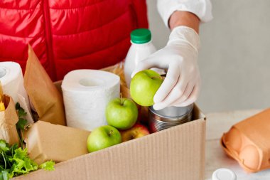 Koruyucu tıbbi maske ve eldiven giyen gönüllü bağış kutusuna yiyecek koyuyor. Kırmızı yelek giyen teslimatçı eleman yiyecek dolu bir kutuyla. Servis karantina pandemik koronavirüsü.