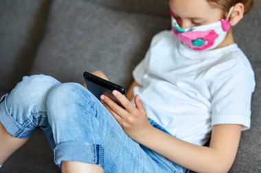 Korunma maskesi takan küçük bir kız evdeki kanepede video oyunu oynuyor, tablet oynuyor ya da öğreniyor, evde oturuyor, Coronavirus karantinası ve sosyal mesafeyi takip ediyor..