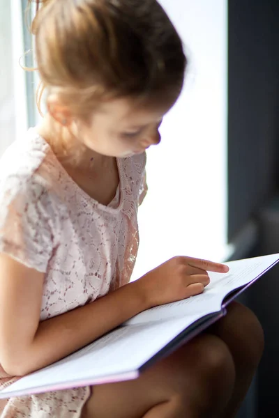Linda niña leyendo libro en casa, en alféizar de la ventana —  Fotos de Stock