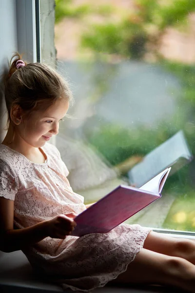 Linda niña leyendo libro en casa, en alféizar de la ventana —  Fotos de Stock