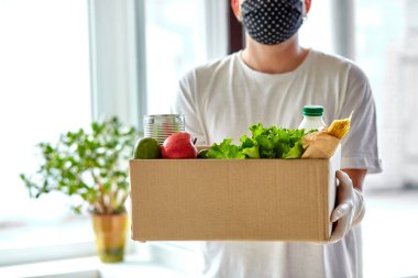 Beyaz koruyucu maskeli ve eldivenli gönüllü eve bağış kutusu getiriyor. Paket kutusu yiyecek taşıyan kurye. Teslimatsız teslimat. Servis karantina pandemik koronavirüsü.