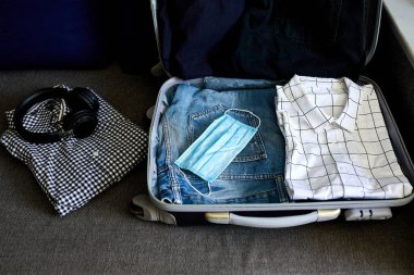 Evde sıradan erkek eşyalarıyla bavul hazırlamak, seyahat, seyahat ve tatil için salgın sırasında koruyucu maske takmak. Boşluğu kopyala.