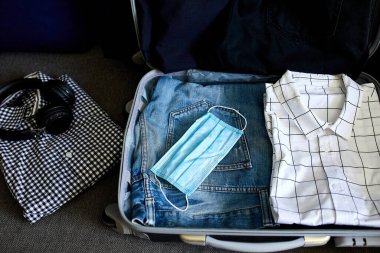 Evde sıradan erkek eşyalarıyla bavul hazırlamak, seyahat, seyahat ve tatil için salgın sırasında koruyucu maske takmak. Boşluğu kopyala.