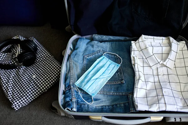 Evde sıradan erkek eşyalarıyla bavul hazırlamak, seyahat, seyahat ve tatil için salgın sırasında koruyucu maske takmak. Boşluğu kopyala.