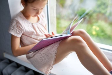 Evde, pencere kenarında kitap okuyan sevimli küçük bir kız evde ders çalışan bir çocuk, karantina, evde kalmak, yaşam tarzı....