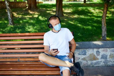 Kulaklıkla müzik dinleyen, parkın dışında yüz koruyucu maske takan, akıllı telefon kullanan, yeni yaşam tarzı, karantina, koronavirüs....