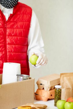 Koruyucu tıbbi maske ve eldiven giyen gönüllü bağış kutusuna yiyecek koyuyor. Kırmızı yelek giyen teslimatçı eleman yiyecek dolu bir kutuyla. Servis karantina pandemik koronavirüsü.