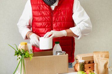 Koruyucu tıbbi maske ve eldiven giyen gönüllü bağış kutusuna yiyecek koyuyor. Kırmızı yelek giyen teslimatçı eleman yiyecek dolu bir kutuyla. Servis karantina pandemik koronavirüsü.