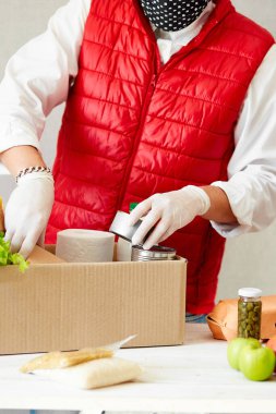 Koruyucu tıbbi maske ve eldiven giyen gönüllü bağış kutusuna yiyecek koyuyor. Kırmızı yelek giyen teslimatçı eleman yiyecek dolu bir kutuyla. Servis karantina pandemik koronavirüsü.