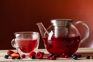 Orman meyveleri çift cam ve cam çaydanlık kırmızı bir arka plan üzerinde ahşap bir masa üzerinde bir fincan çay nane.