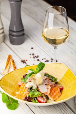 Sezar salatası somon. karışımı salata, domates, kaşar peyniri, fesleğen. Bir seramik tabak bir tabak bir restoranda ahşap bir masa var. Bir kadeh beyaz şarap tablodur. seçici odak