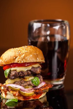 Amerikan fast food konsepti. Sulu bir Amerikan hamburgeri, iki biftek köftesi ve cam bir masada koyu bir bira ve turuncu bir arka plan. Boşluğu kopyala