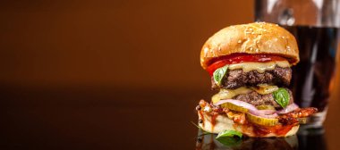 Amerikan fast food konsepti. Sulu bir Amerikan hamburgeri, iki biftek köftesi ve cam bir masada koyu bir bira ve turuncu bir arka plan. Boşluğu kopyala