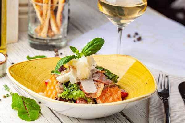 Caesar salad with salmon. mix of salads, cherry tomatoes, parmesan cheese, basil. A dish in a ceramic plate is on a wooden table in a restaurant. A glass of white wine is on the table. selective focus