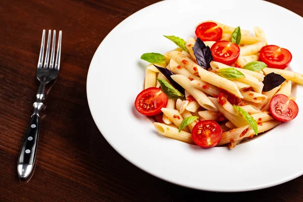 Domates ve fesleğen çatal yanındaki ahşap bir masa üzerinde bir beyaz forvor plaka ile domates soslu penne makarna. Arka plan görüntüsü. İtalyan makarna tarifleri için kavramı. kopya alanı, üstten görünüm