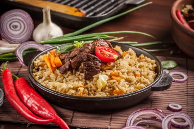 Doğu, özbek mutfağı kavramı. Biber pilav, plov bir siyah dökme demir tavada yalan malzemeler, soğan, domates, maydanoz, yanındaki ahşap bir masa üzerinde. Orta Asya mutfağı.