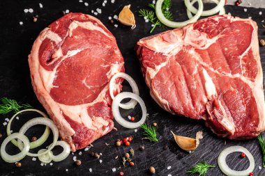 Çiğ et T-bone biftek bir parçası üzerinde siyah bir seramik baharatlarla taş yatıyor. soğan, maydanoz ve sarımsak. Görünümü üst