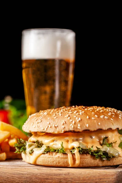 The concept of American cuisine. Juicy burger with meat patty, with cheese and french fries lying on a wooden board in a restaurant. Near a glass of beer with foam.