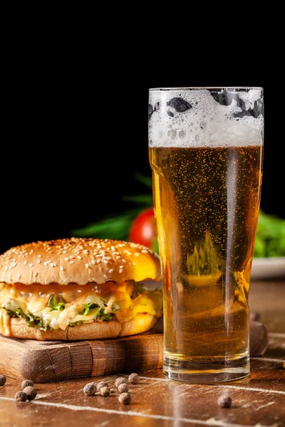 The concept of American cuisine. Juicy burger with meat patty, with cheese and french fries lying on a wooden board in a restaurant. Near a glass of beer with foam.