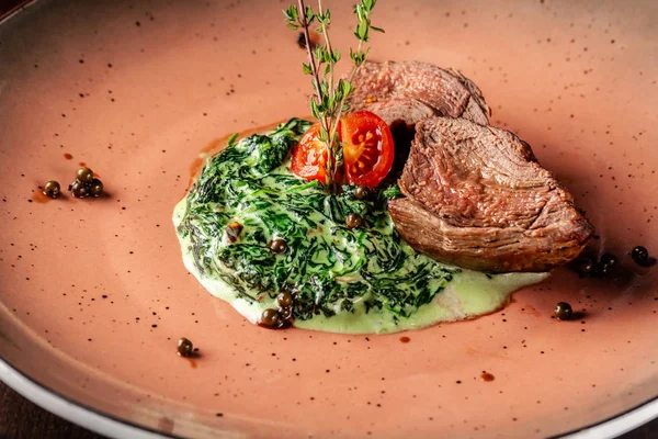 The concept of Italian cuisine. Juicy veal steak on spinach pillow with cream, cherry tomatoes and thyme. Next to the table is a glass of red wine. Beautiful serving dishes in the restaurant.