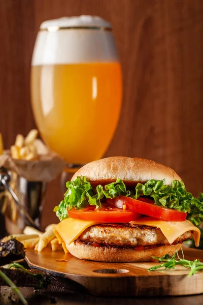 American cuisine concept. Juicy burger with meat patty, tomatoes, cheddar cheese, lettuce and homemade bun. In the background are french fries and a glass of beer. Close up