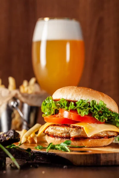 American cuisine concept. Juicy burger with meat patty, tomatoes, cheddar cheese, lettuce and homemade bun. In the background are french fries and a glass of beer. Close up