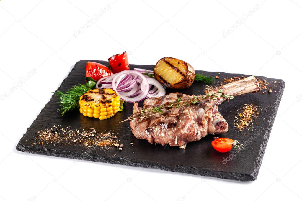 Large meat steak on the bone, grilled, served with grilled vegetables, corn, red onion, sweet peppers, potatoes. Modern serving dishes in the restaurant on a black slate blackboard.