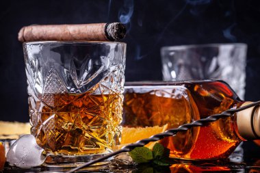 alkolizm kavramı, yalnızlık, bağımlılık. Bir şişe viski, bir bardak viski ve buzlu, siyah bir masada dumanlı bir puro, siyah bir arka plan üzerinde. Erkeksi rahatlama için elit içecek