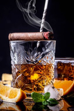 alkolizm kavramı, yalnızlık, bağımlılık. Bir şişe viski, bir bardak viski ve buzlu, siyah bir masada dumanlı bir puro, siyah bir arka plan üzerinde. Erkeksi rahatlama için elit içecek