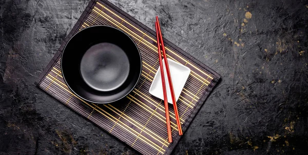 Empty Japanese dishes. A black ceramic bowl for Chinese noodles or Thai soup lies on a bamkuk rug. White saucepot for soy sauce and red Chinese sticks on a black background. Top view, copy space