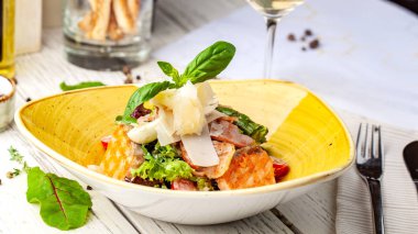 Sezar salatası somon. karışımı salata, domates, kaşar peyniri, fesleğen. Bir seramik tabak bir tabak bir restoranda ahşap bir masa var. Bir kadeh beyaz şarap tablodur. seçici odak