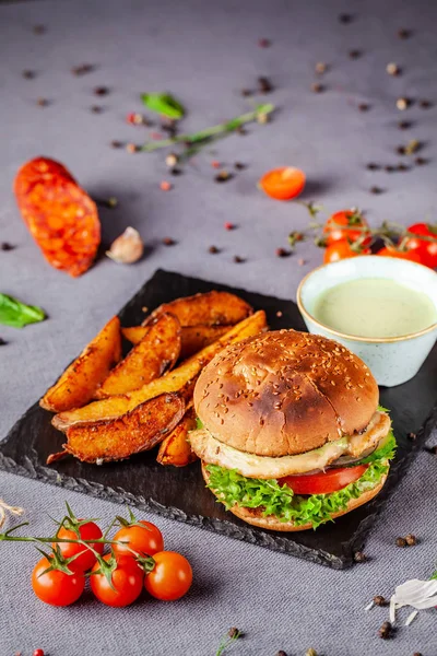 American burger with chicken cutlet, tomato, cucumber, green sauce. Sauce and for french fries on a black stone slate board. Dish in a fast food restaurant. Copy space