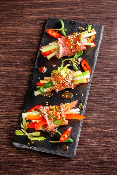 Concept of European cuisine. Snack for wine. Raw veal roll with crispy vegetables, cucumber, celery, red bell pepper. Serving dishes in the restaurant on a black stone slate board