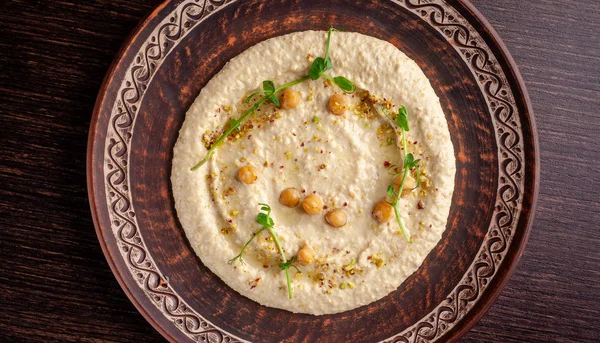 The concept of Arabic cuisine. Oriental hummus with grilled sesame and pistachios. A dish in a restaurant in a red ceramic dish on a wooden table. Top view, copy space