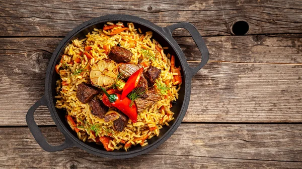 O conceito de cozinha oriental. Pilaf uzbeque nacional com carne em uma frigideira de ferro fundido, em uma mesa de madeira. imagem de fundo. vista superior, espaço de cópia, leigos planos — Fotografia de Stock